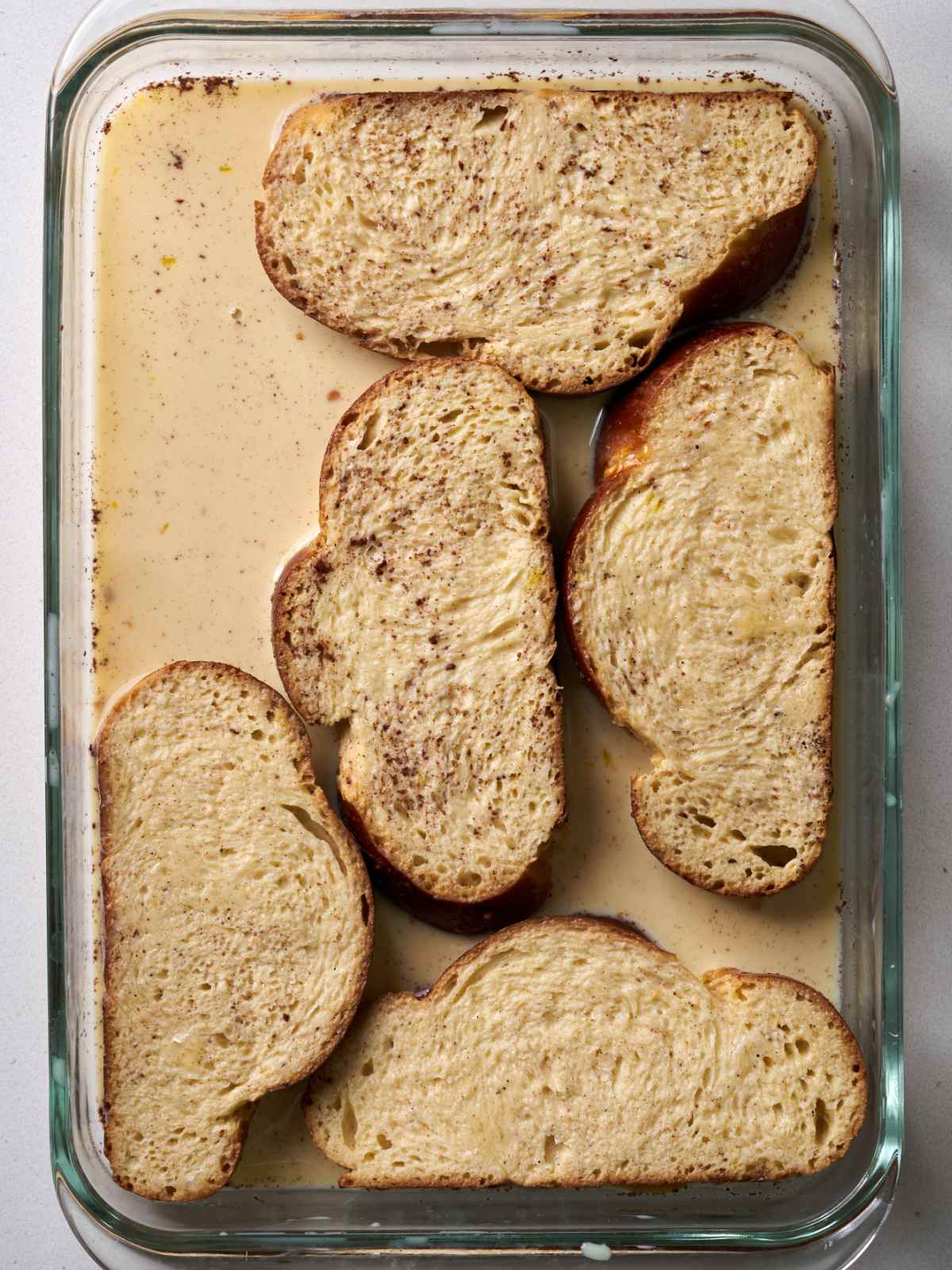 A large glass baking dish with five slices of challah bread soaking in a light brown liquid.