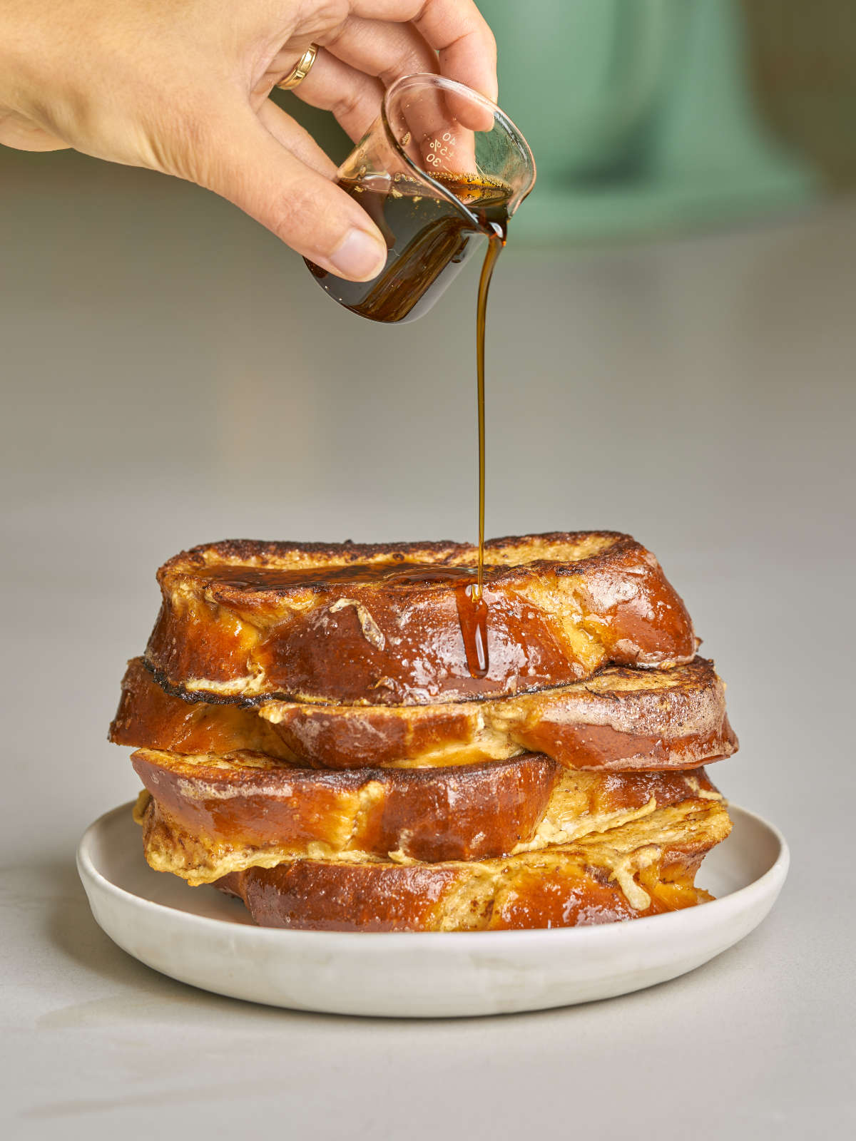 A woman's hand drizzling maple syrup from a small glass beaker onto a stack of challah French toast.