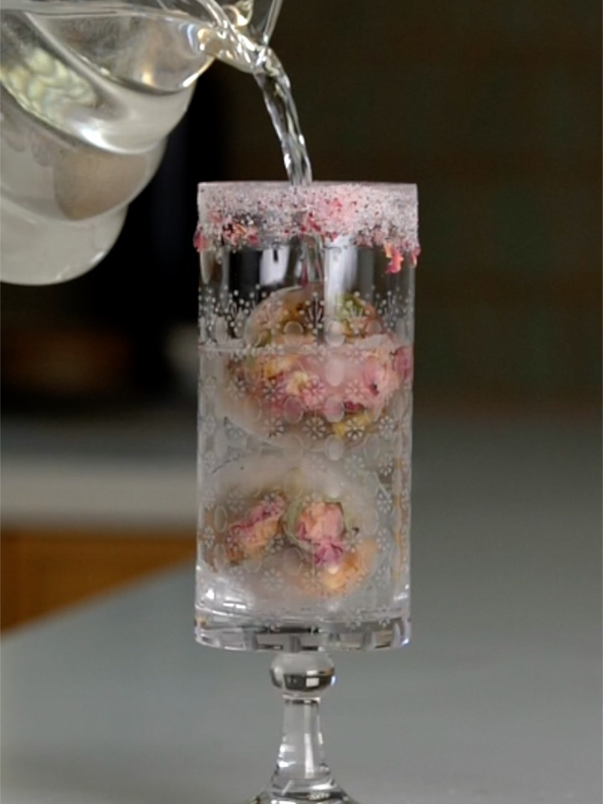 A glass pitcher pouring water into a tall patterned glass with pink sugar rim and roce ice cubes.