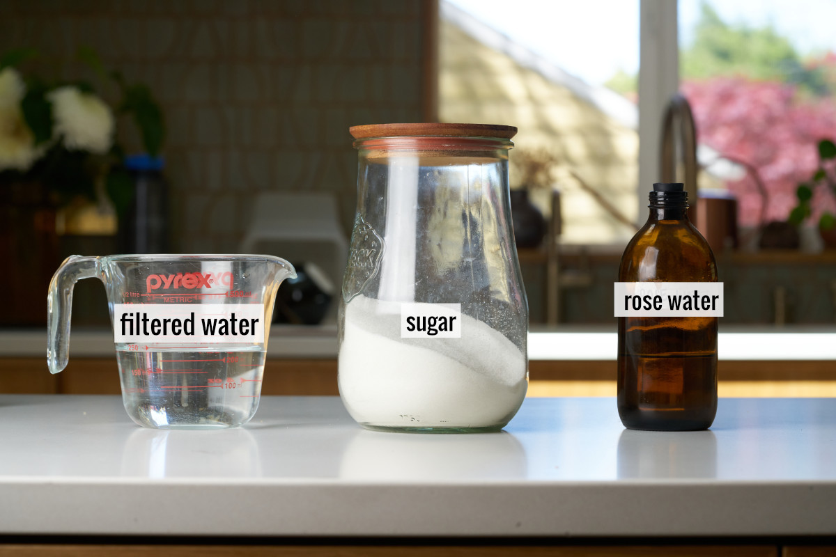 A liquid measuring cup with water next to a container with sugar and an amber bottle with rose water.