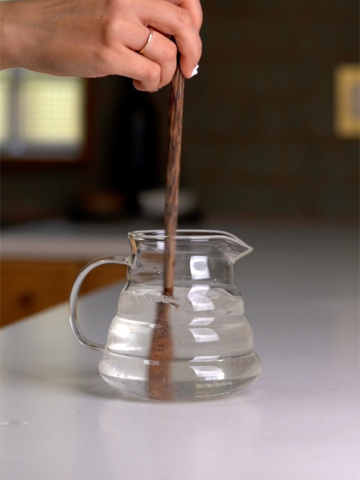 A hand using a long wooden spoon to stir the liquid in a glass measuring jug on a marble counter.