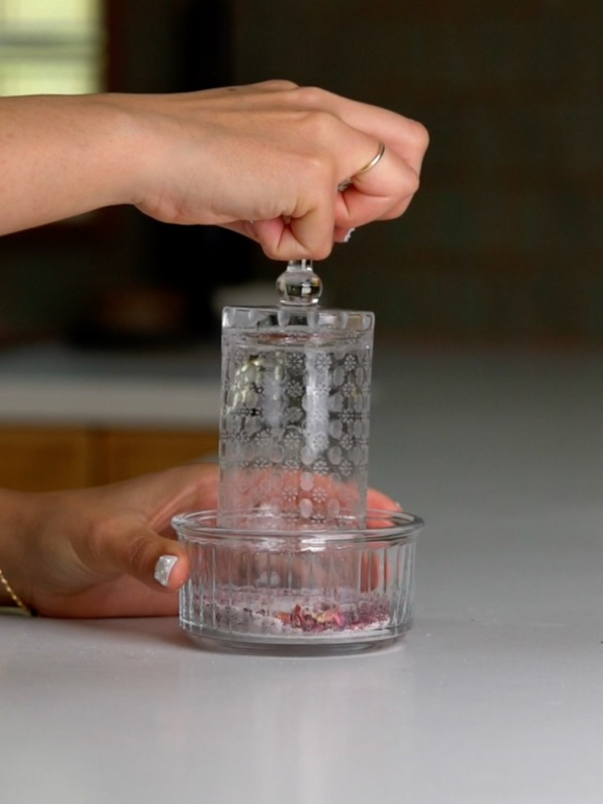 A hand dipping a tall patterned glass into a glass dish with pink sugar.