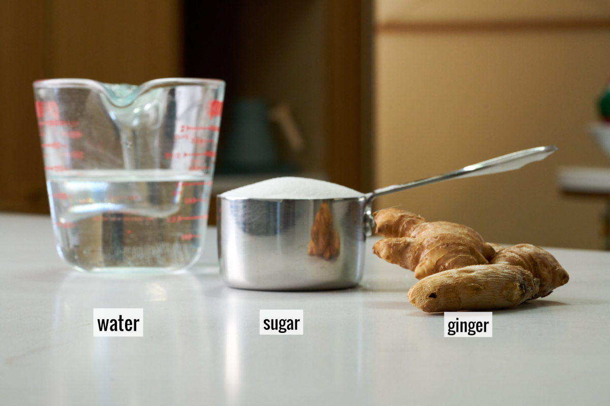 Liquid measuring cup with water next to a metal measuring cup with sugar and a knob of ginger.