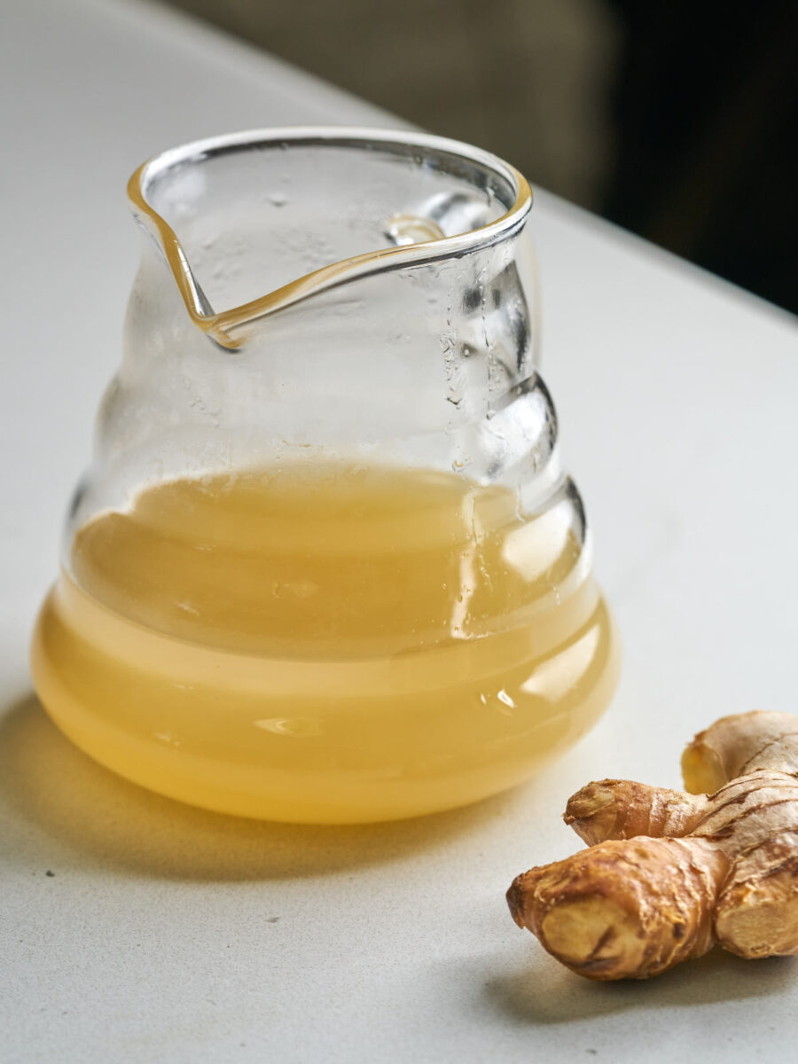 Glass pitcher filled part way with yellow juice next to a knob of ginger.