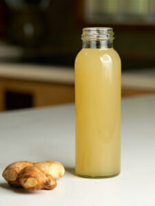 Bottle of yellow juice in a cylindrical glass bottle next to a knob of ginger.
