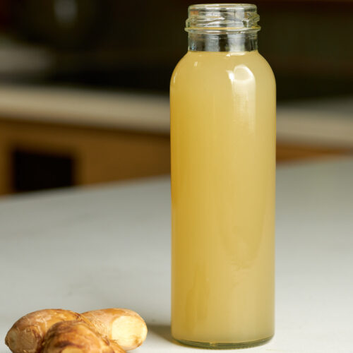 Bottle of yellow juice in a cylindrical glass bottle next to a knob of ginger.