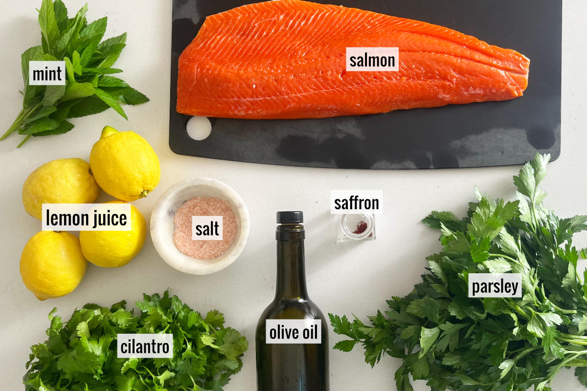 Ingredients to make lemon herb salmon including lemons, herbs, and salmon next to a bottle of olive oil and a dish of pink salt.