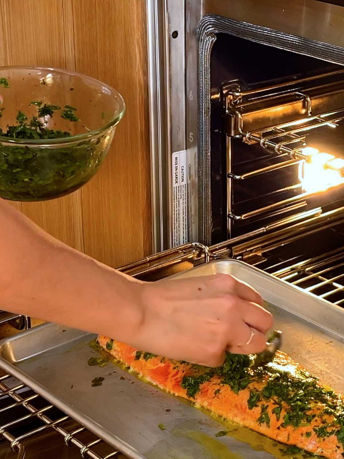 Baking sheet with salmon on an oven rack with a hand pouring herb sauce on top of the salmon filet.