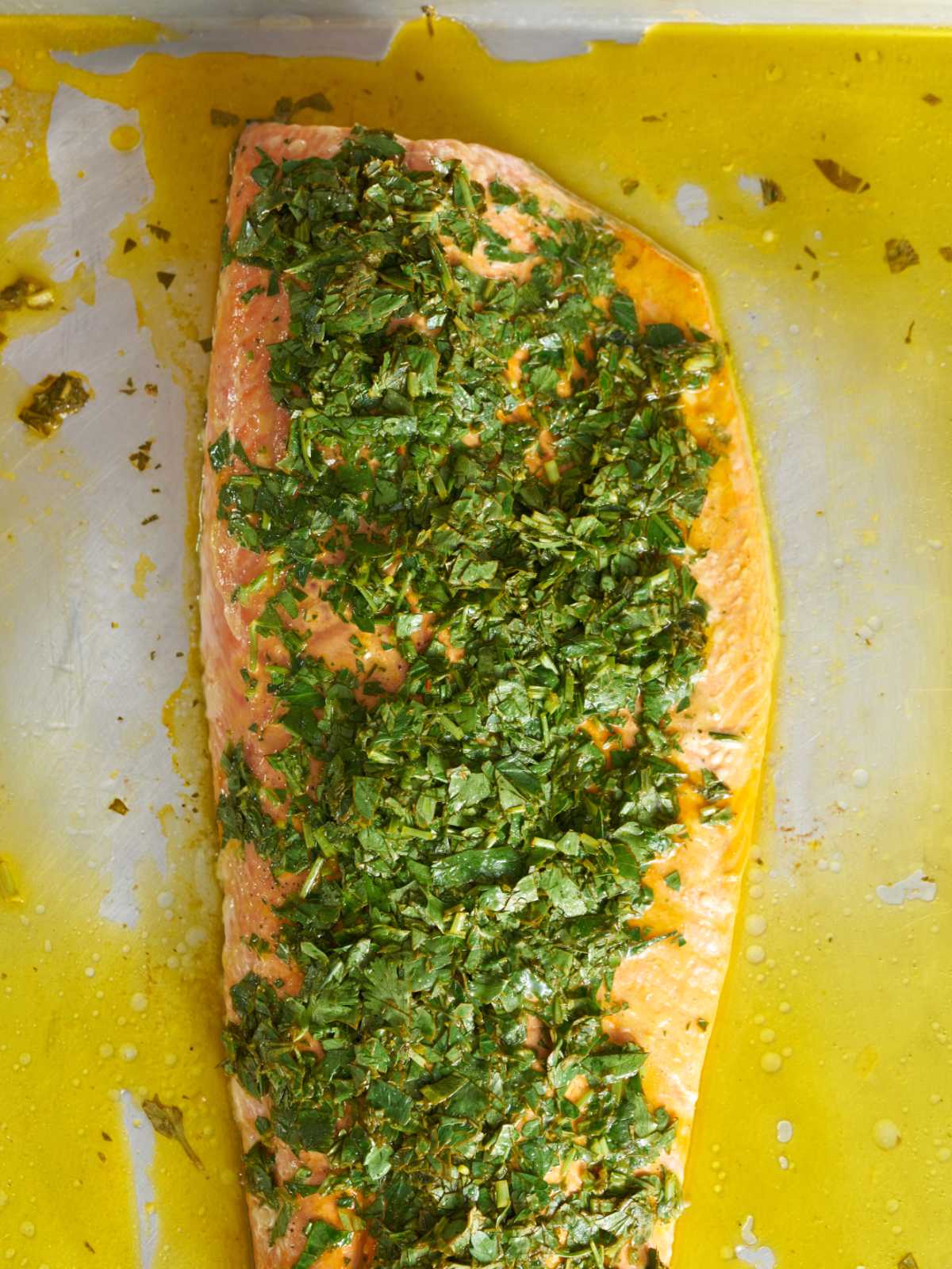 Cooked salmon on a baking sheet covered in herbs and surrounded by yellow sauce.