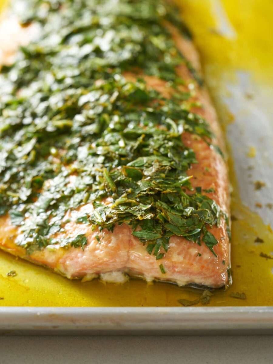 Front view of a salmon filet covered in herbs and surrounded by yellow sauce on a baking sheet.