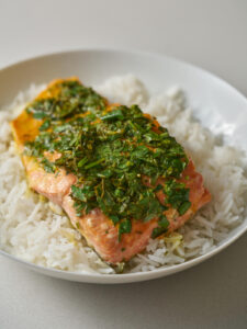 A portion of salmon covered in herbs and on a bed of white rice in a white pasta dish.