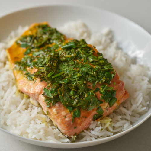 A portion of salmon covered in herbs and on a bed of white rice in a white pasta dish.