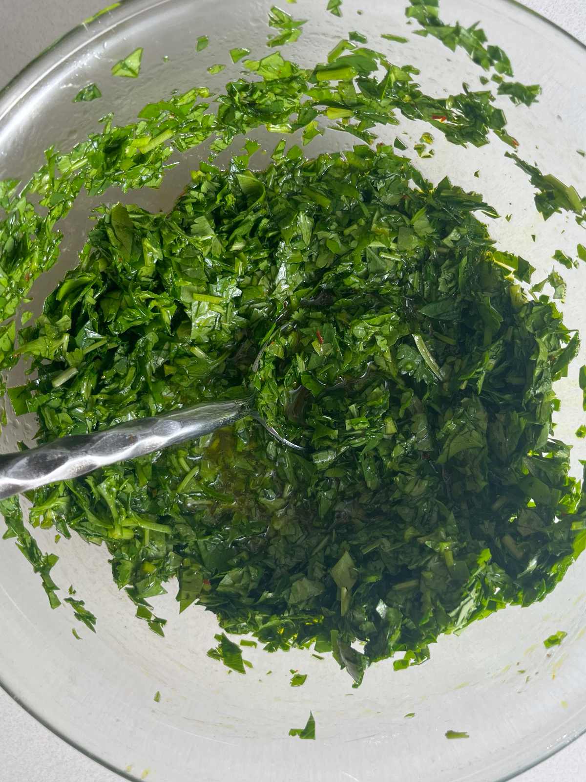 Chopped herbs in a glass bowl with a metal spoon.