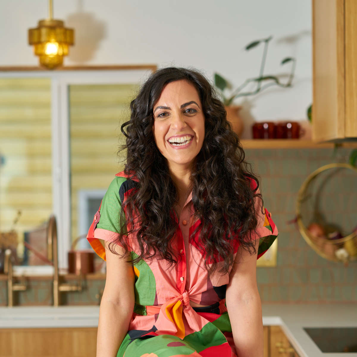 Woman sitting on a kitchen countertop with brown curly hair and a colorful jumpsuit.