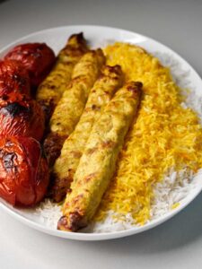Four ground chicken kabobs next to four barbecued tomatoes on a bed of yellow rice.