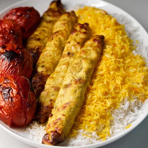 Four ground chicken kabobs next to four barbecued tomatoes on a bed of yellow rice.