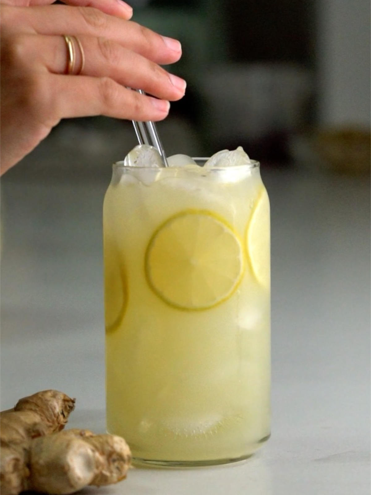 Hand holding a glass straw in a tall glass of lemonade with lemon slices next to a knob of ginger.