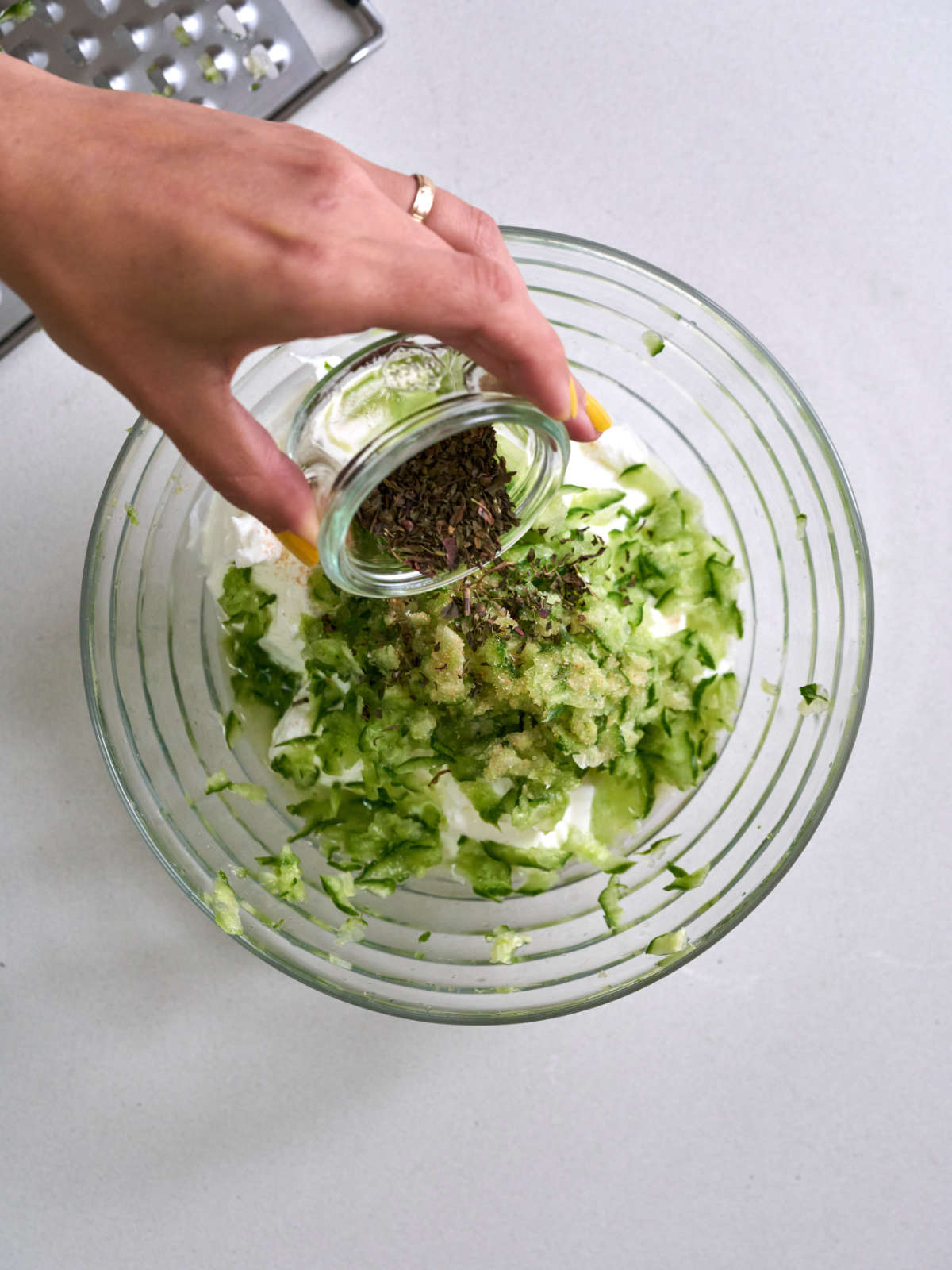 Hand pouring dried herbs into a glass bowl with grated cucumber.