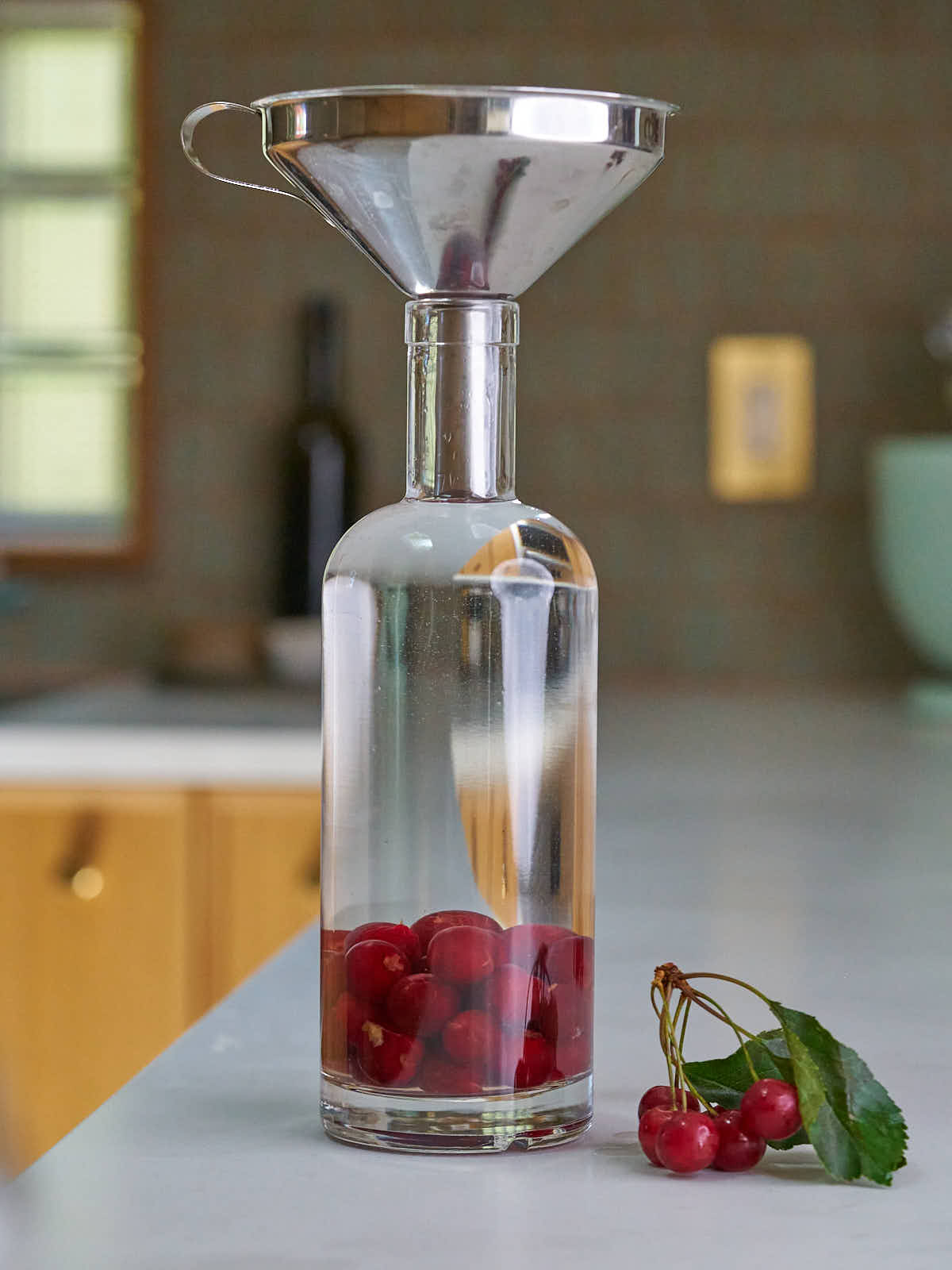 Bottle with cherries and clear liquid next to a bunch of cherries and topped with a metal funnel.