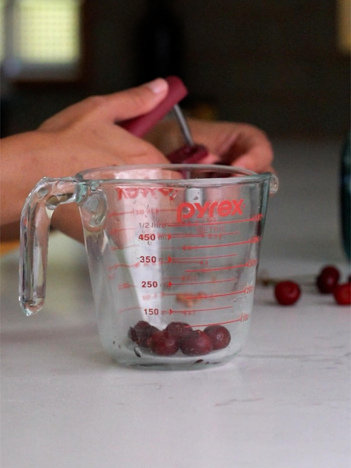 Pitting cherries behind a liquid measuring cup with a few cherries inside.