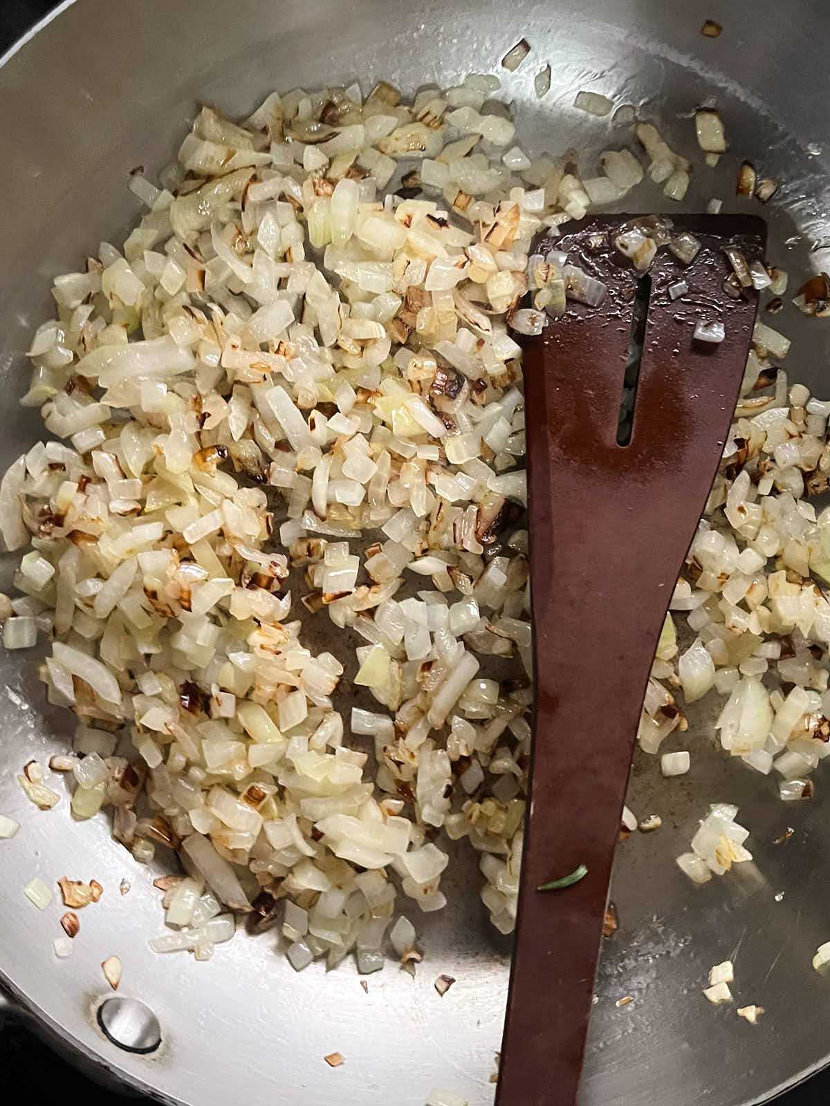 Onions and spices cooking in a large silver pan with a wooden spoon.