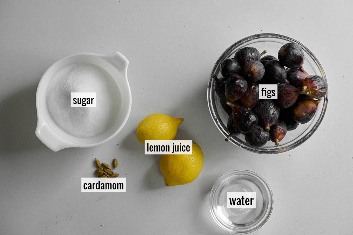 Figs in a bowl next to two lemons, a bowl of sugar, cardamom pods and a small bowl of water.