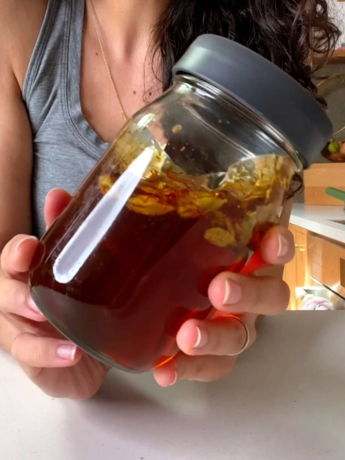 Two hands holding a jar of honey with cardamom pods in it and a dark grey lid.