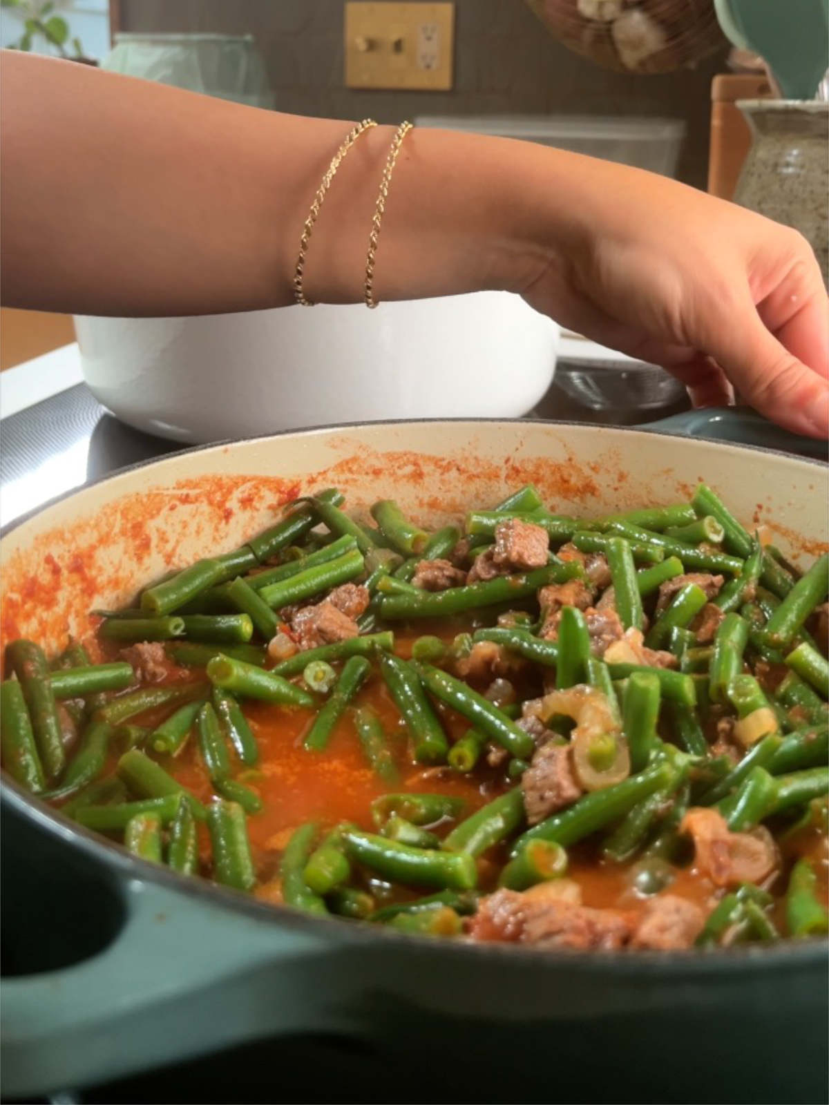 Hands grabbing a pot on a stove with green beans and tomato sauce in it.