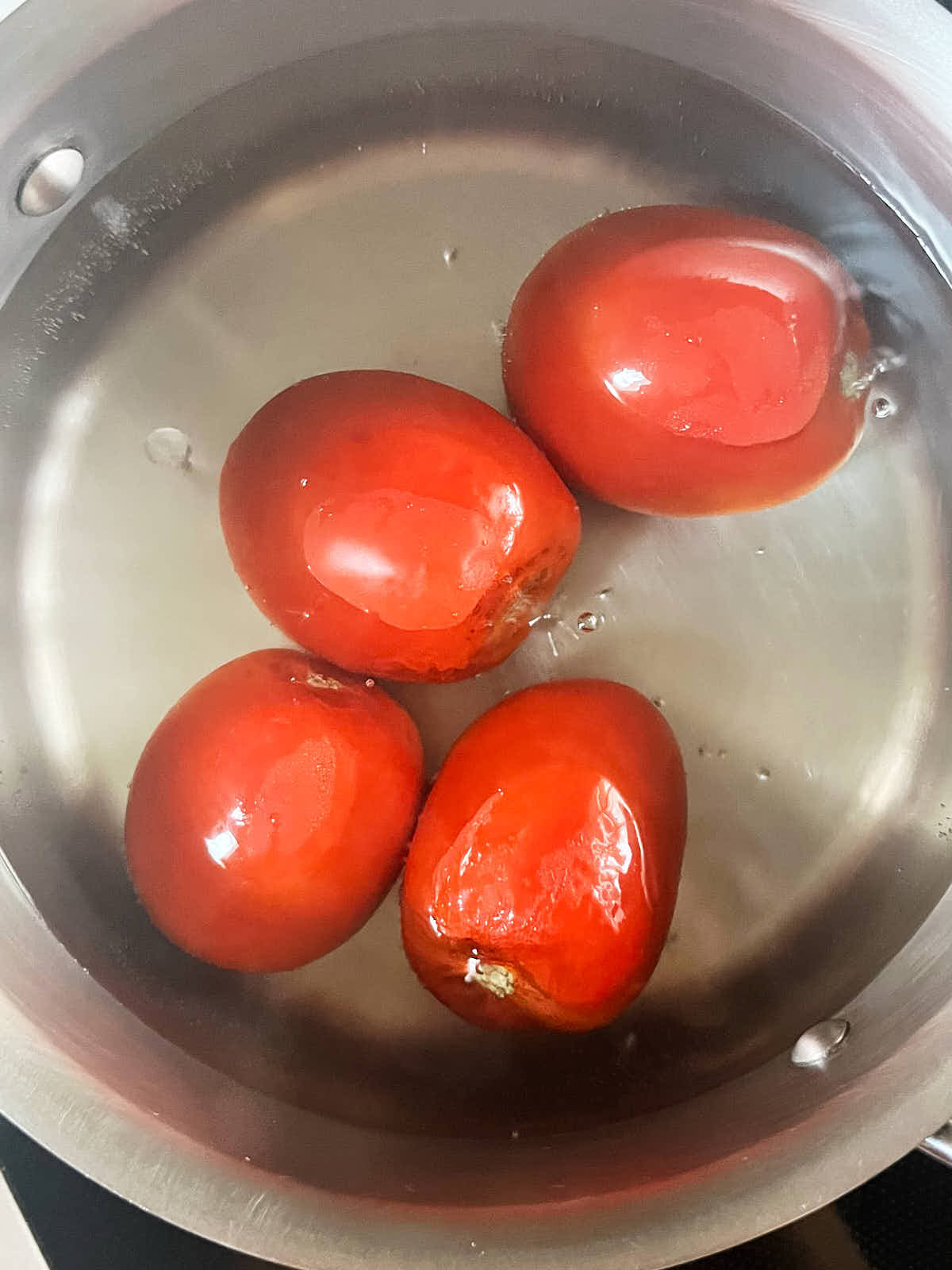 Four tomatoes in a metal pot filled with simmering water.