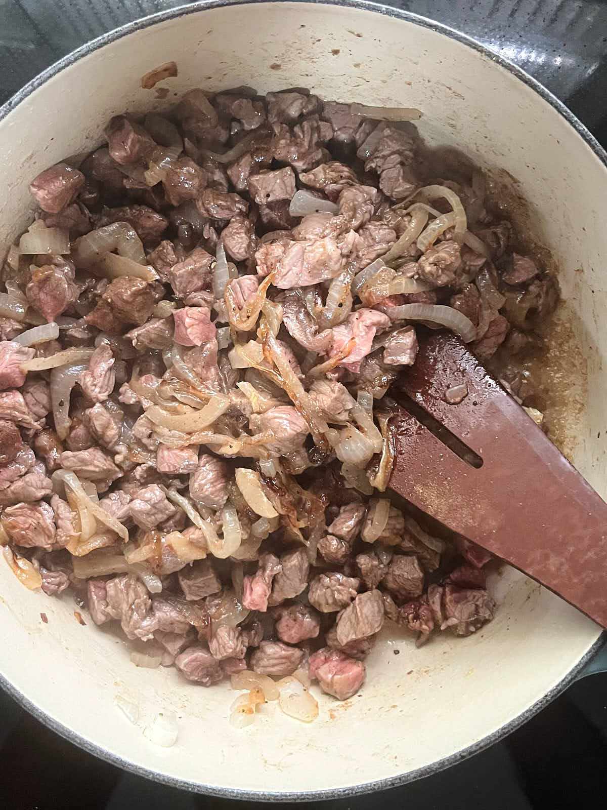 Small cubes of meat and sauteed onions in an enameled cast iron pot with a wooden utensil.