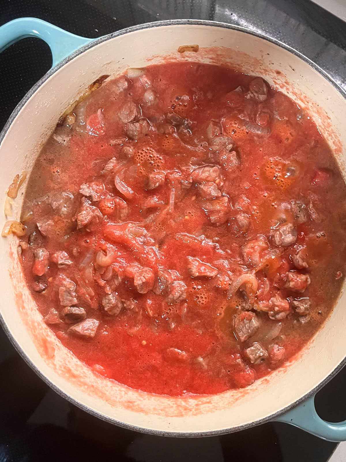 Meat cubes in tomato sauce in an enameled cast iron pan.