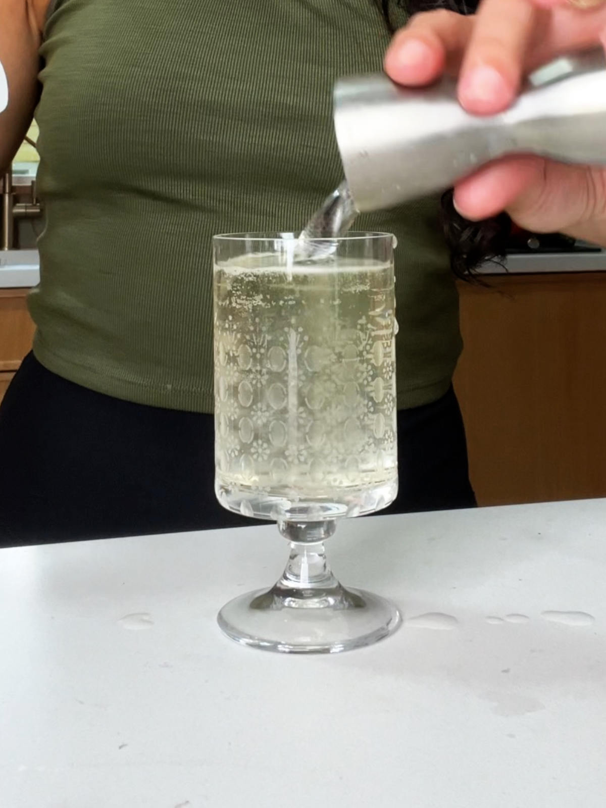 Woman in a green shirt and black bottoms pouring liquid into a glass pedestal glass from a jigger.