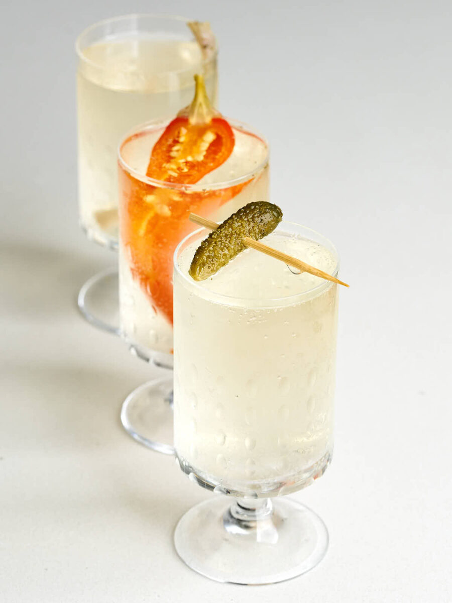 Small pedestal glass with off-white bubbly liquid and a cornichon garnish in front of another with half a red pepper.