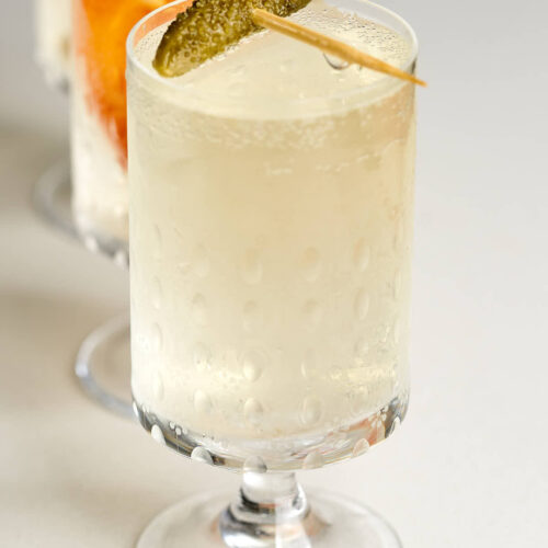 Small pedestal glass with off-white bubbly liquid and a cornichon garnish.