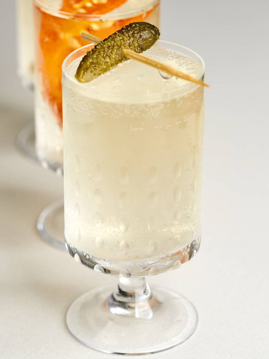 Small pedestal glass with off-white bubbly liquid and a cornichon garnish.