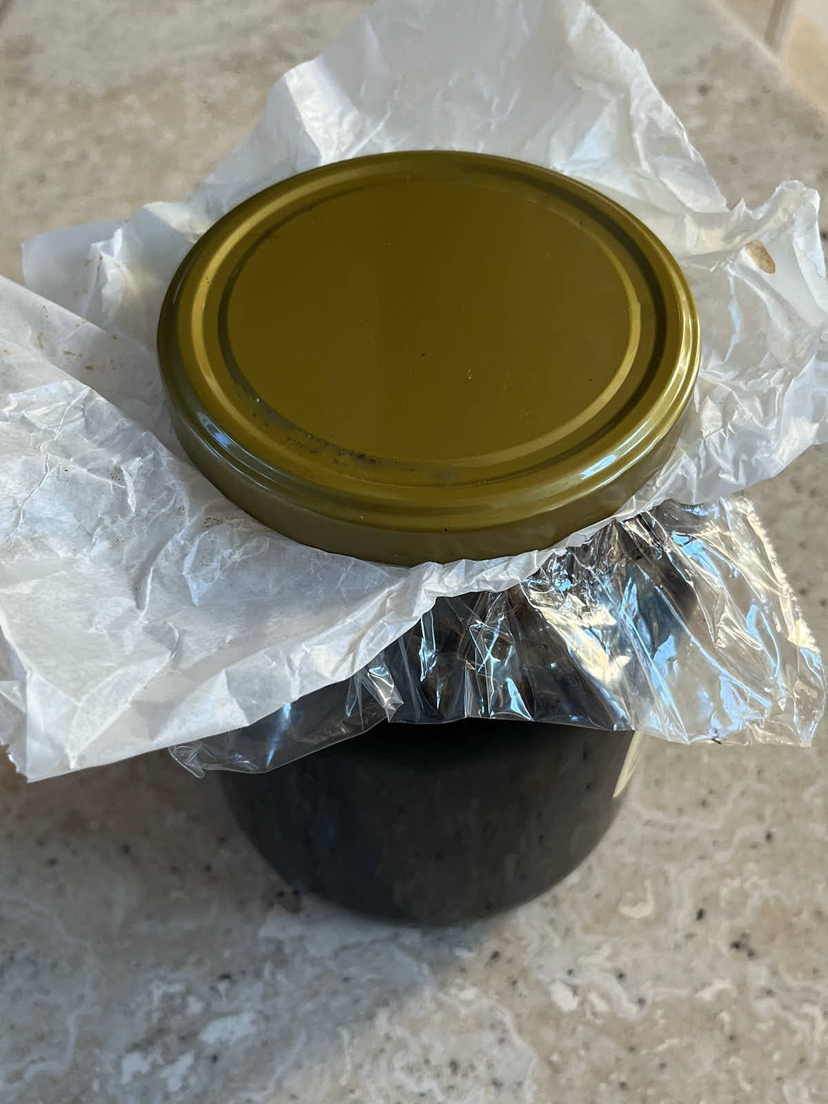 Jar of black contents with a gold metal lid and a layer of parchment paper and plastic between the lid and jar.