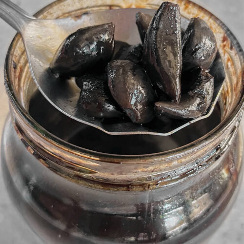 Metal spoon holding black garlic cloves over a jar of black garlic.