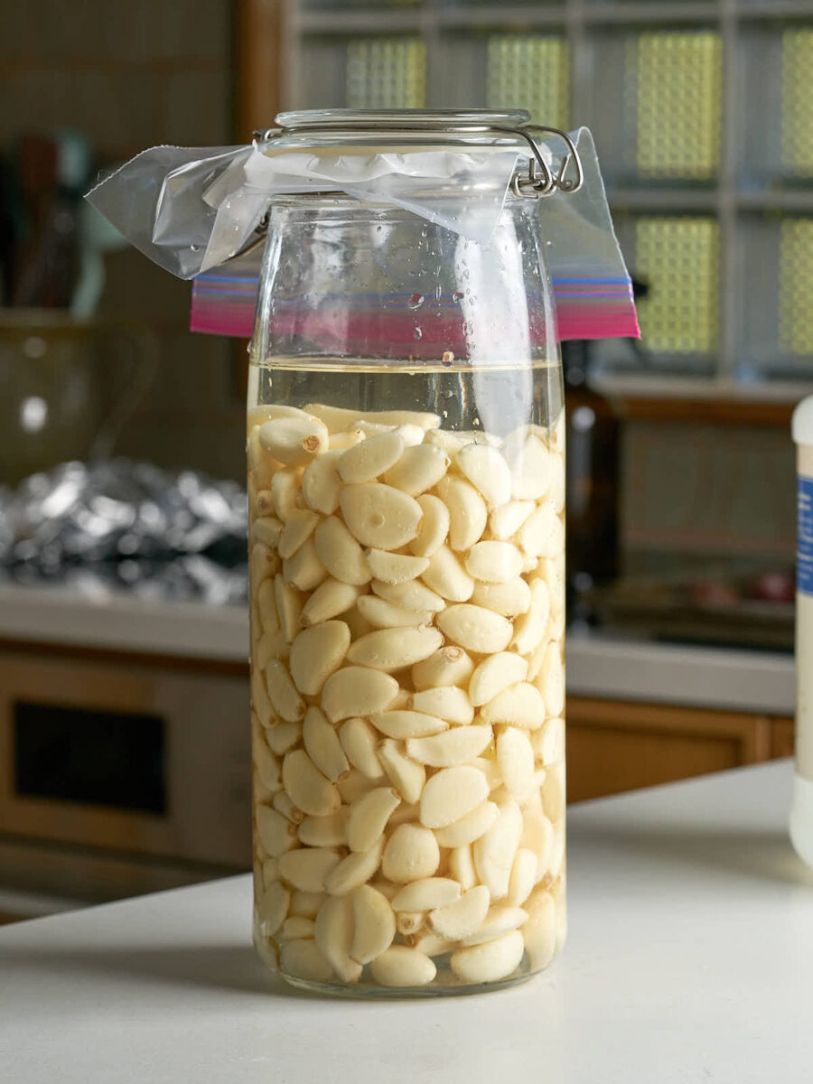 Glass jar filled with peeled garlic cloves topped with clear liquid and sealed with a layer of parchment paper and a plastic bag.