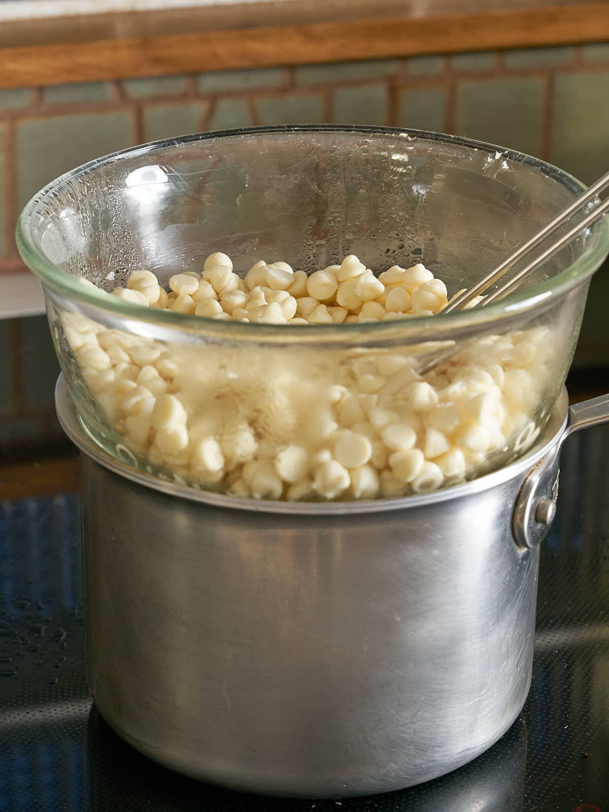 White chocolate chips melting in a glass bowl over a silver pot.