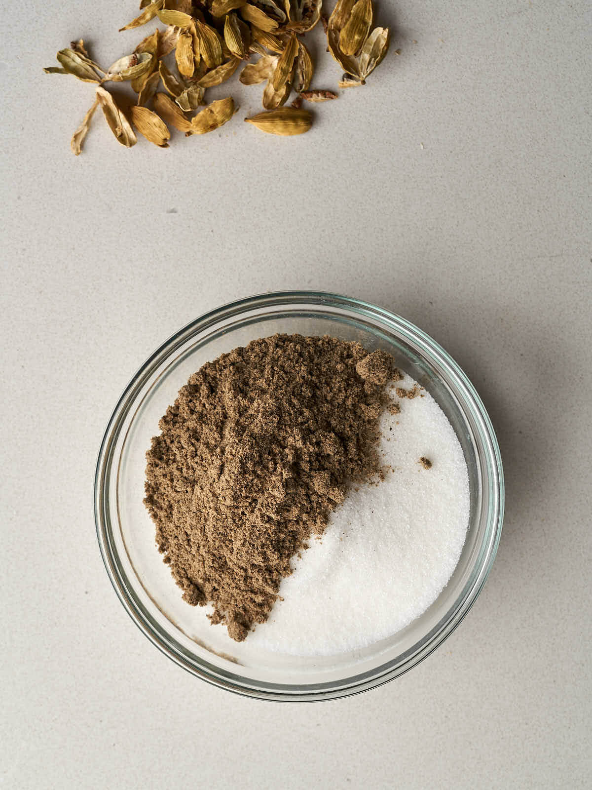 A small glass bowl filled with sugar and topped with ground cardamom next to a pile of cardamom husks.