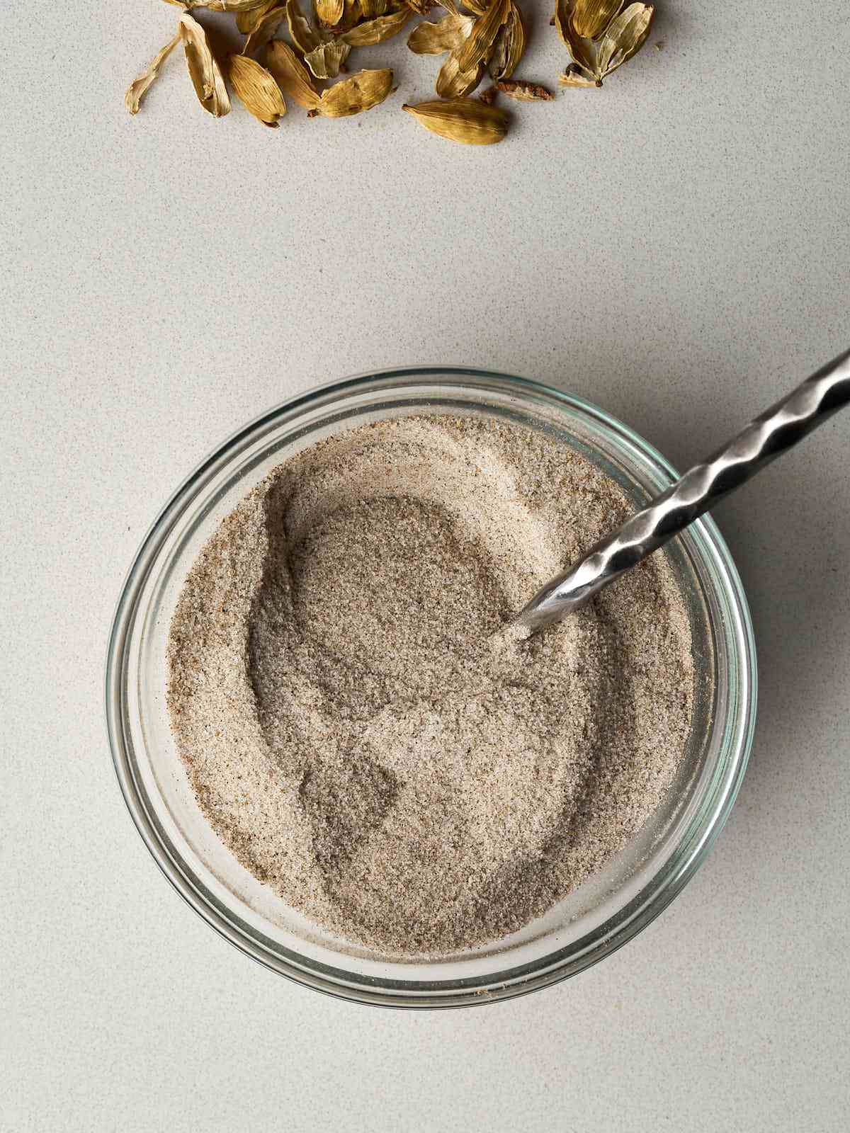 Cardamom and sugar mixed together in a glass bowl with a metal spoon.