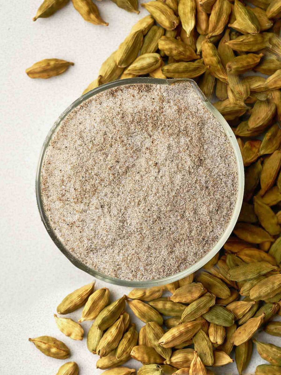 Cardamom sugar in a glass bowl over a pile of cardamom pods.