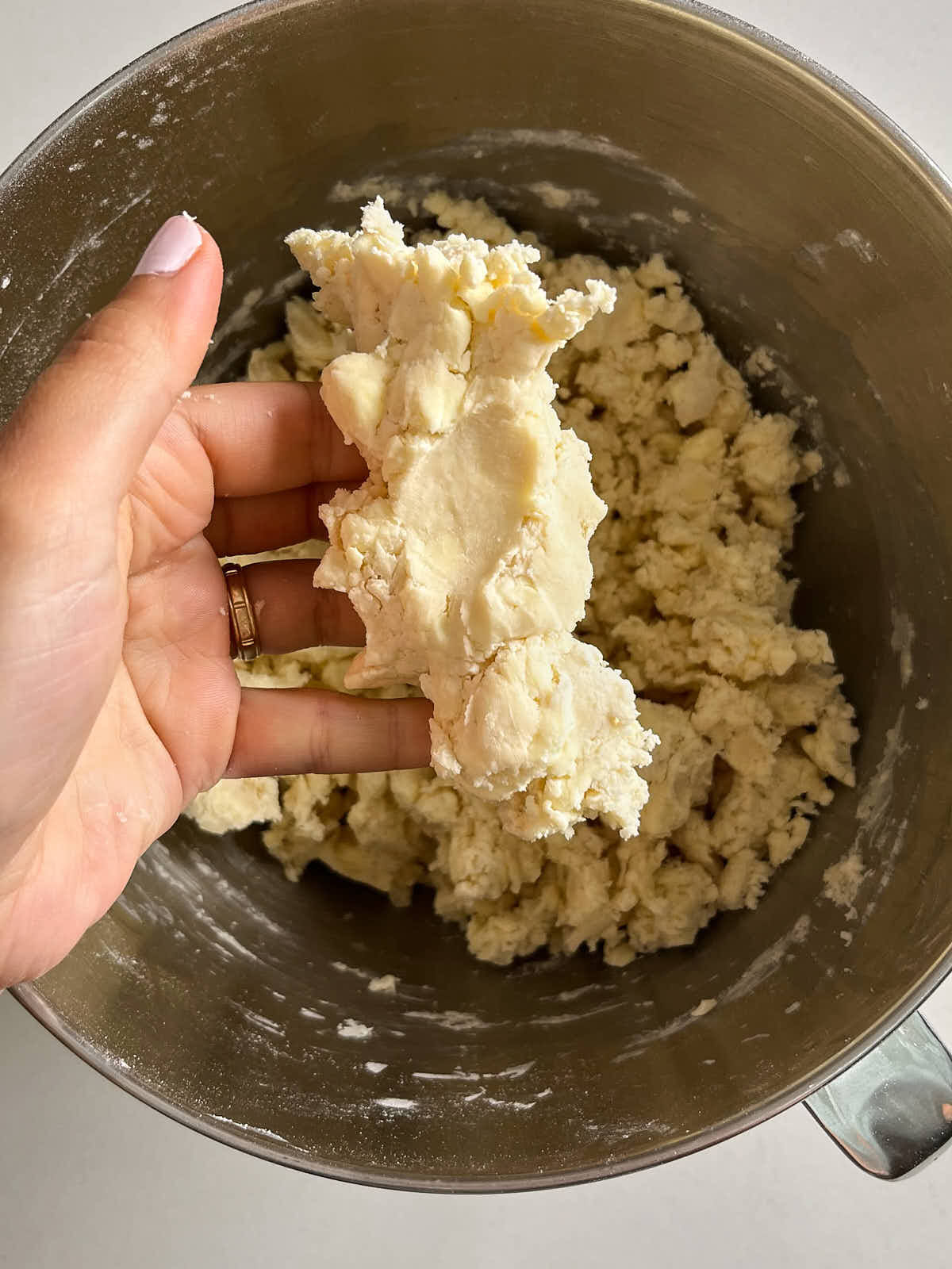 Hand holding a chunk of pastry dough that was squished in the hand over a metal bowl with the rest of the dough.