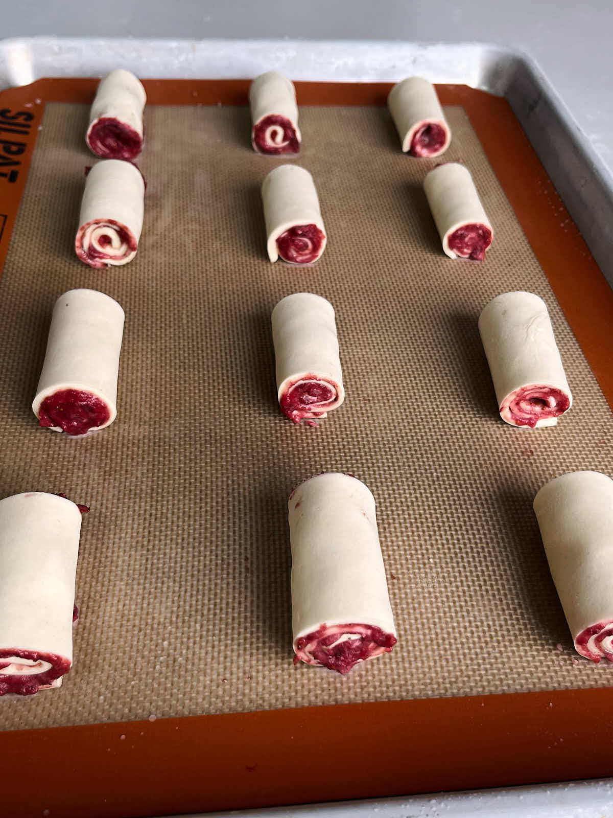 Twelve rolls of pastry dough and red jam arranged on a silicone baking mat.