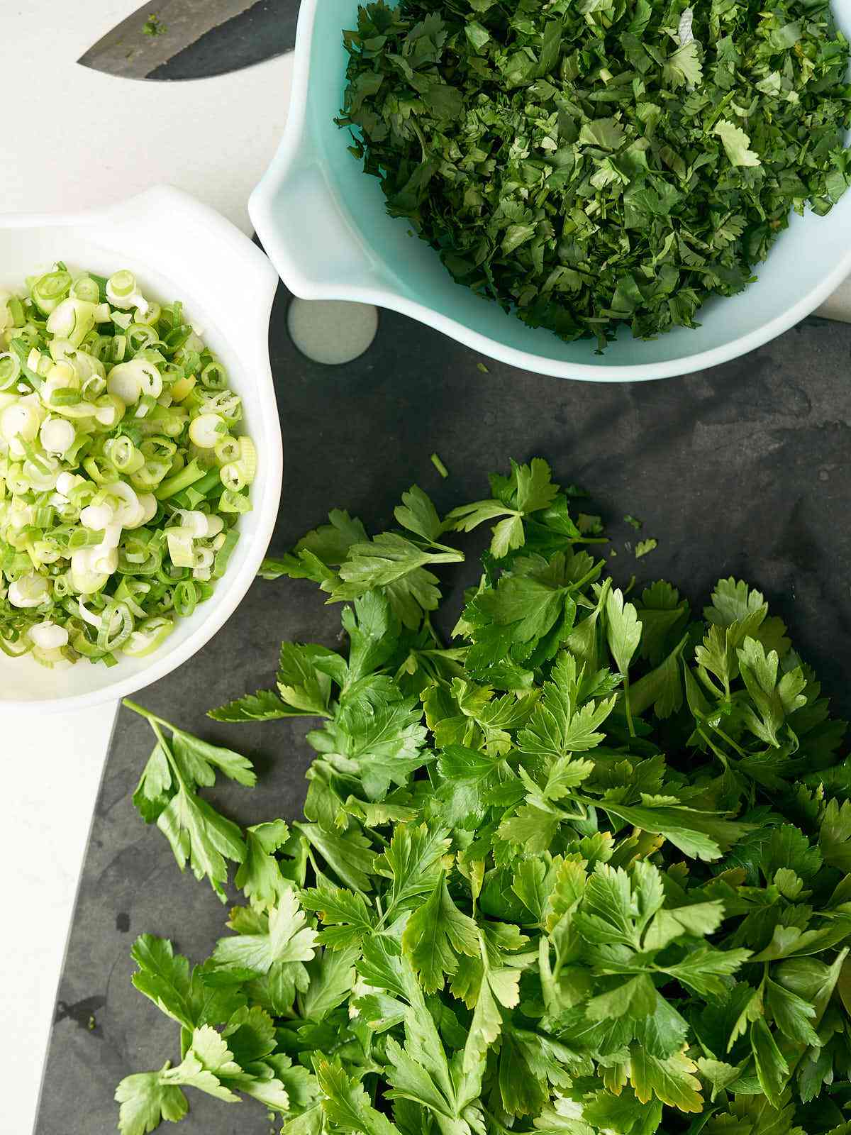 Chopped cilantro in a bowl next to a bowl of green onions and more bunches of cilantro.