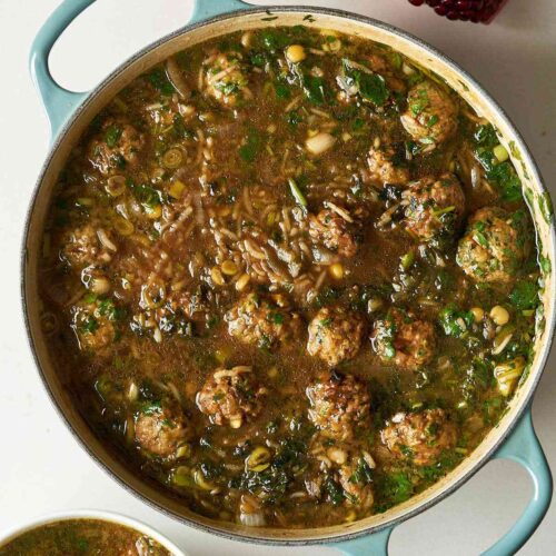 A brown stew with herbs, meatballs, and rice in a large dutch oven with a cut pomegranate beside it.