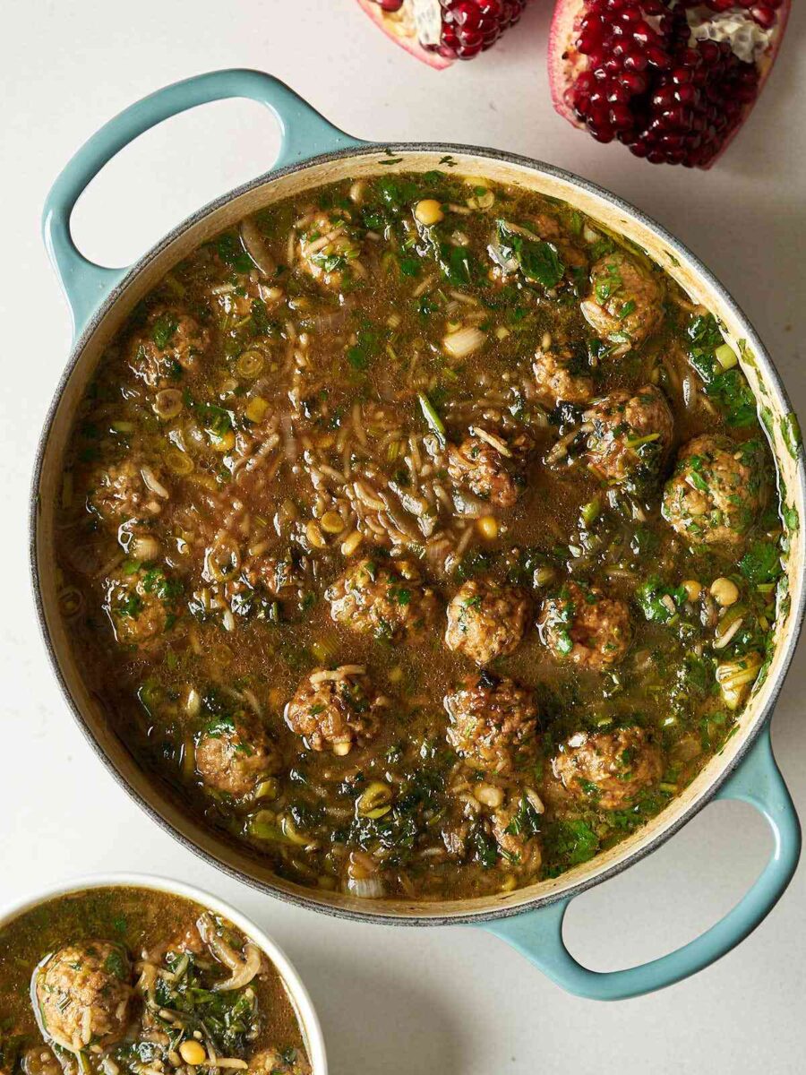 A brown stew with herbs, meatballs, and rice in a large dutch oven with a cut pomegranate beside it.