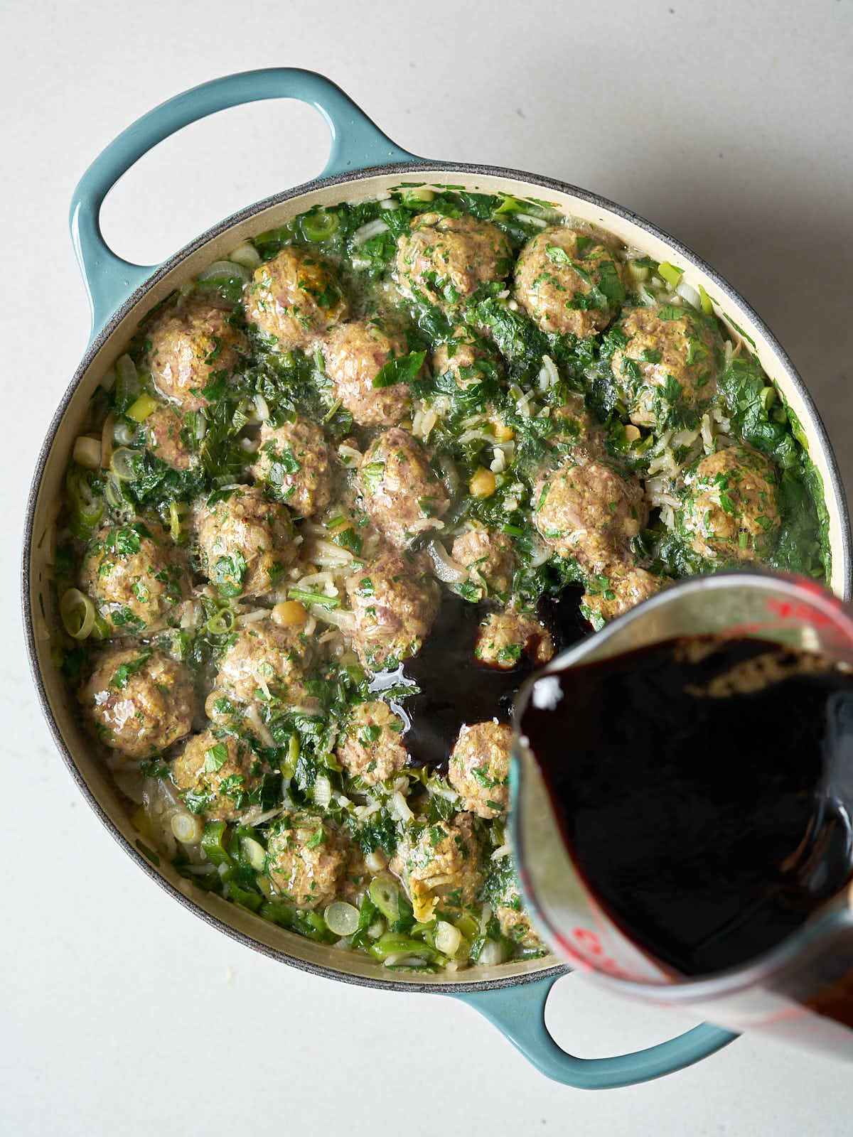 A glass measuring cup pouring a dark liquid into a pot of herbs and meatballs.