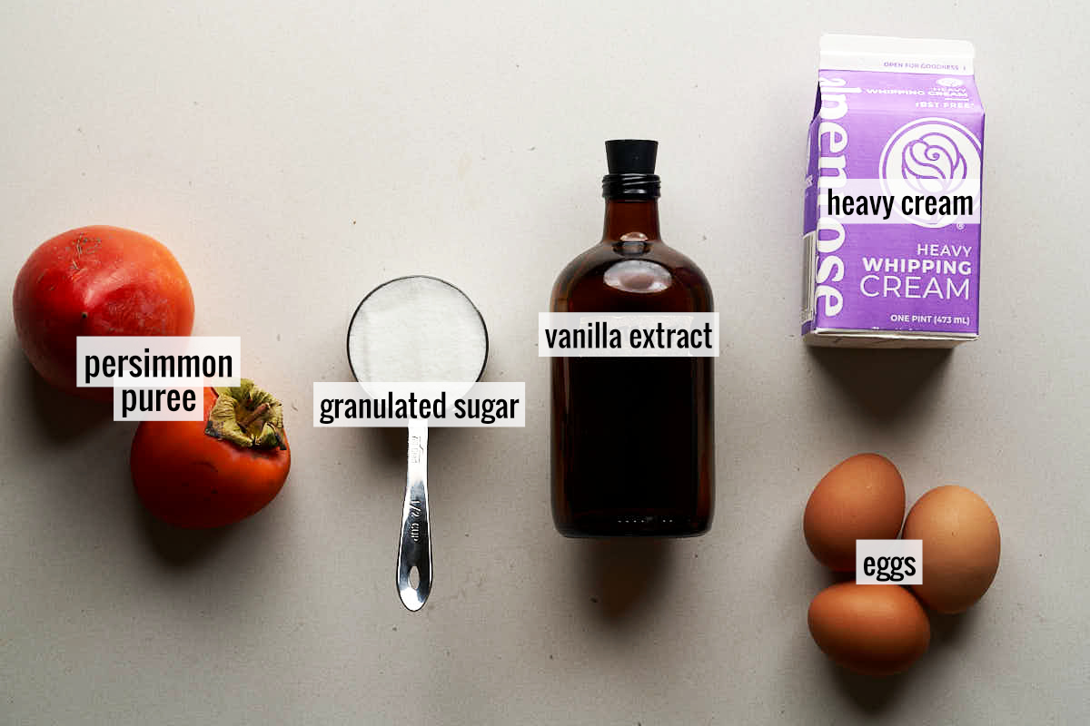 Ingredients to make persimmon semifreddo on a countertop including two persimmons, an amber bottle of vanilla extract, eggs, and sugar in a metal measuring cup.