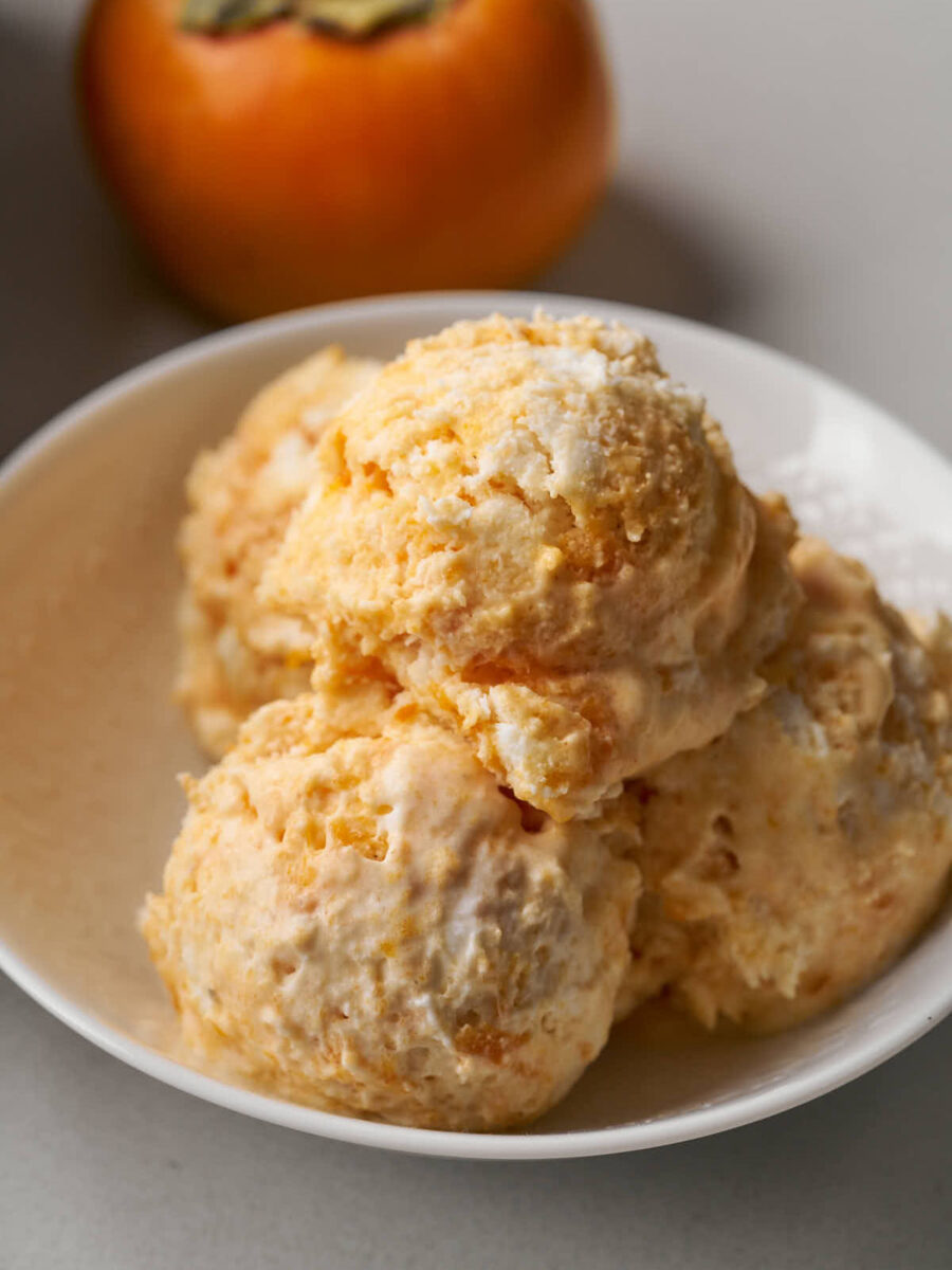 Four scoops of orange semifreddo in a small white bowl next to a persimmon.